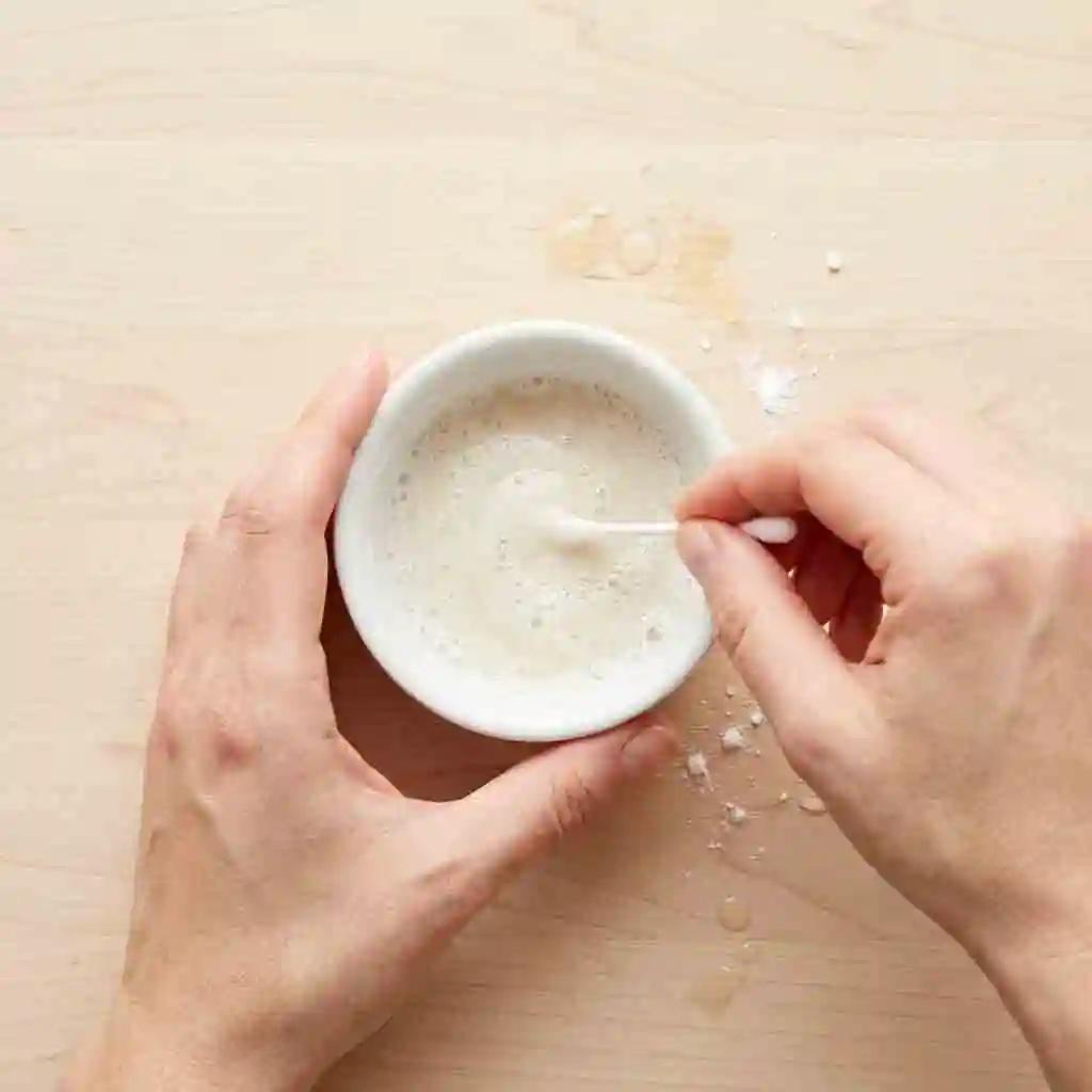 Hands mixing baking soda and dish soap in a small bowl to create a home remedy paste on how to remove beard dye from stained skin.
